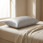 Funda de almohada de seda gris perla sobre una cama de lino hueso junto a la ventana